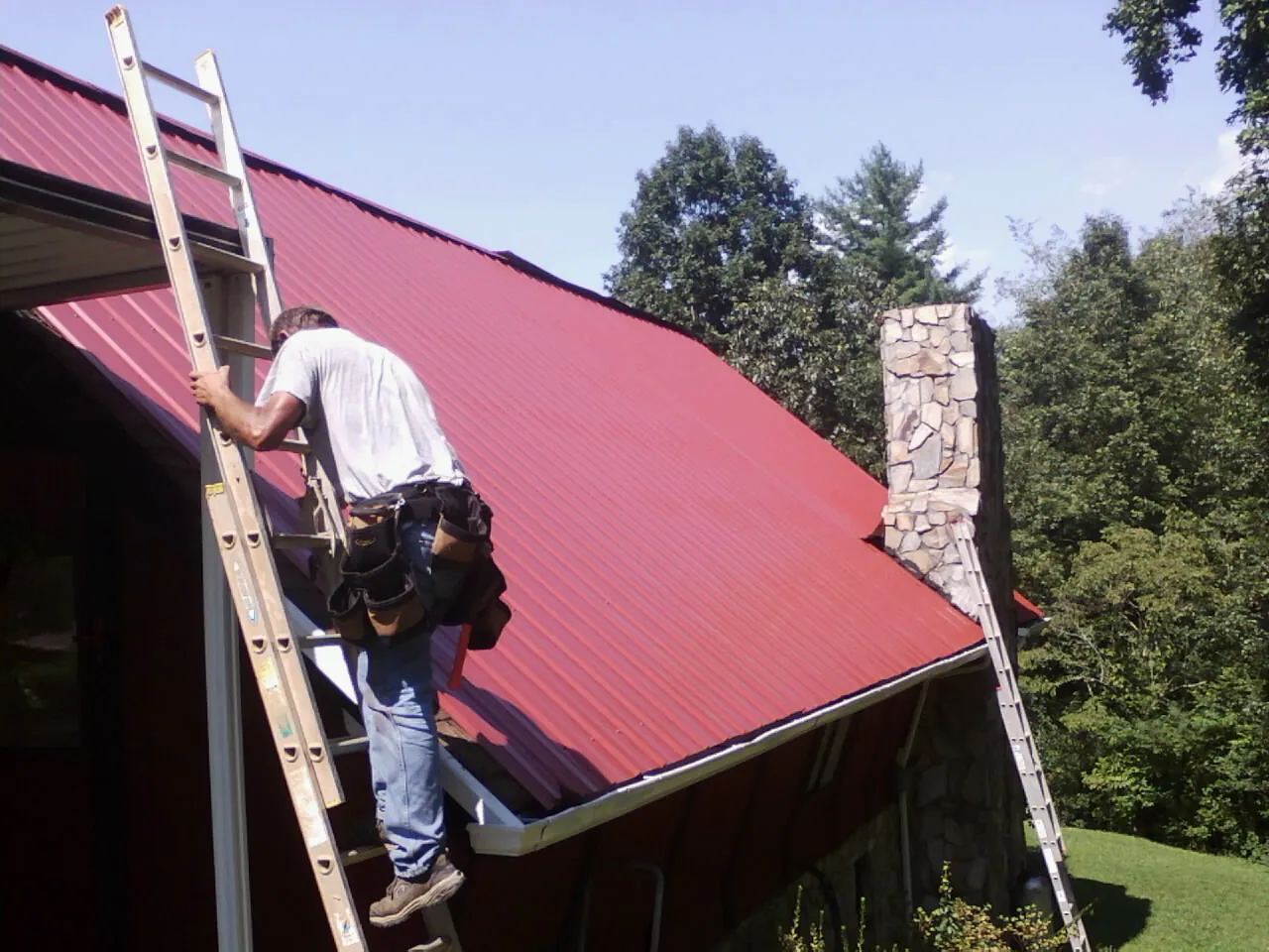 Roofer on a ladder beside a red metal roof and stone chimney — Soffit & Fascia Repair by Roofing Brothers in Rosedale